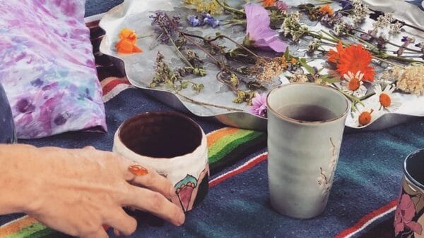 hand thrown ceramic cups with wild foraged tea flowers and leaves nearby, a hand reaches for the steeping wild foraged tea in the ceramic cup