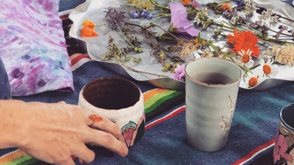 hand thrown ceramic cups with wild foraged tea flowers and leaves nearby, a hand reaches for the steeping wild foraged tea in the ceramic cup