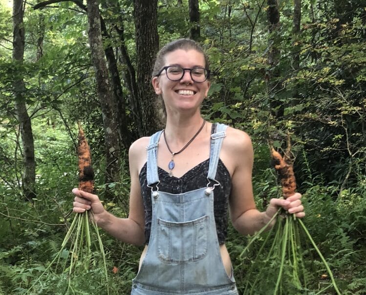 Jenn Tuft, Firefly Gathering Kitchen Director smiles in the garden holding two handfuls of carrots