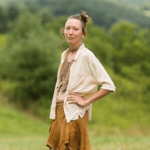 Rain Hall stands in front of a green hill, wearing a buckskin outfit they crafted