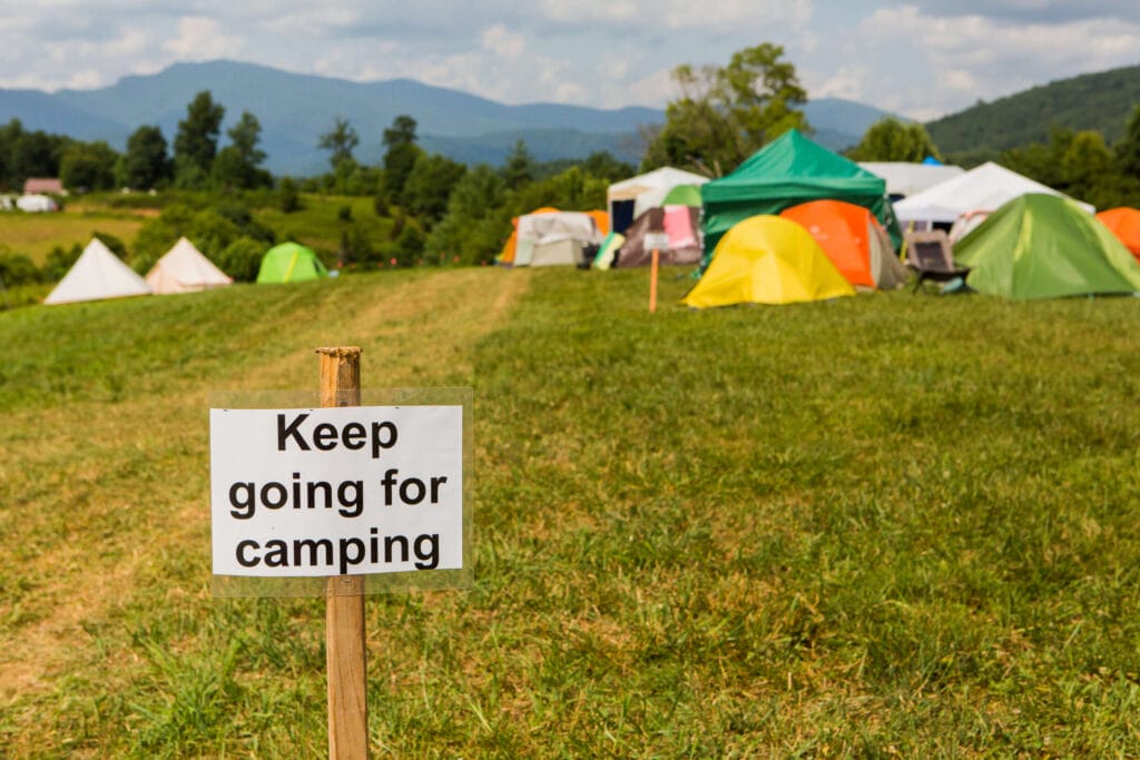 A sign in a field says 'keep going for camping' with tents in the distance