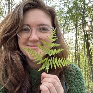 Lark Frazier wears a green sweater and is in a forest in the spring, holding a fern next to her face and smiling