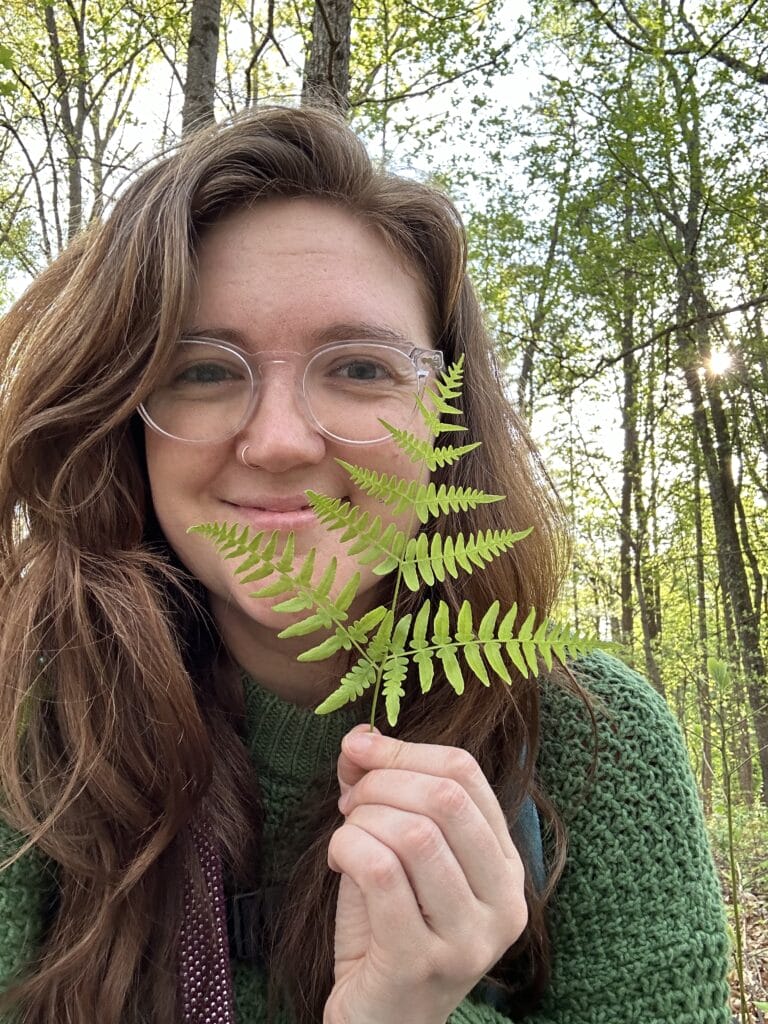 Lark Frazier wears a green sweater and is in a forest in the spring, holding a fern next to her face and smiling