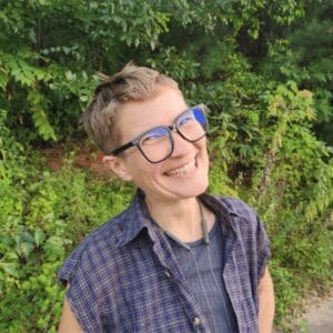 Staj smiles at the camera and is wearing glasses and a plaid shirt