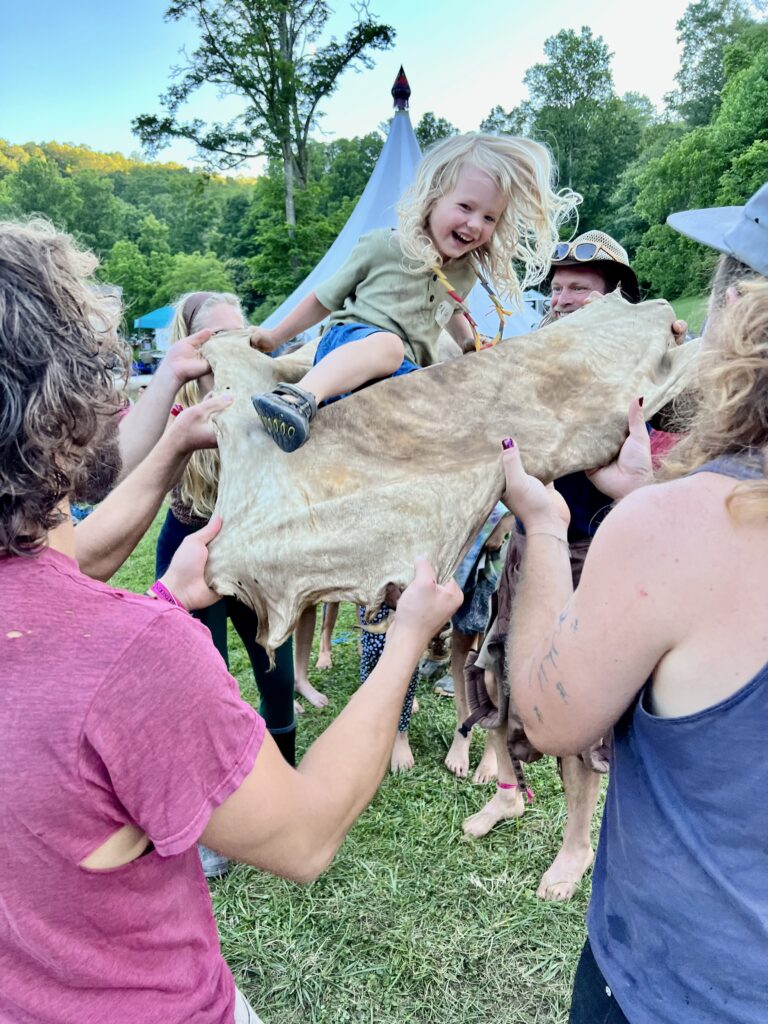 A group of adults lift a small child on a piece of buckskin while the child laughs